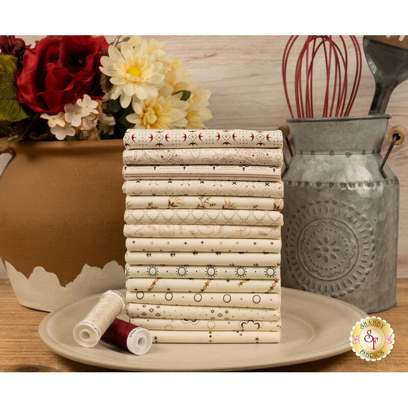 A stack of the fabrics included in the Buttercream & Brickle Bits FQ set, with a jug and a vase and a whisk and spatula and flowers and thread and a plate.