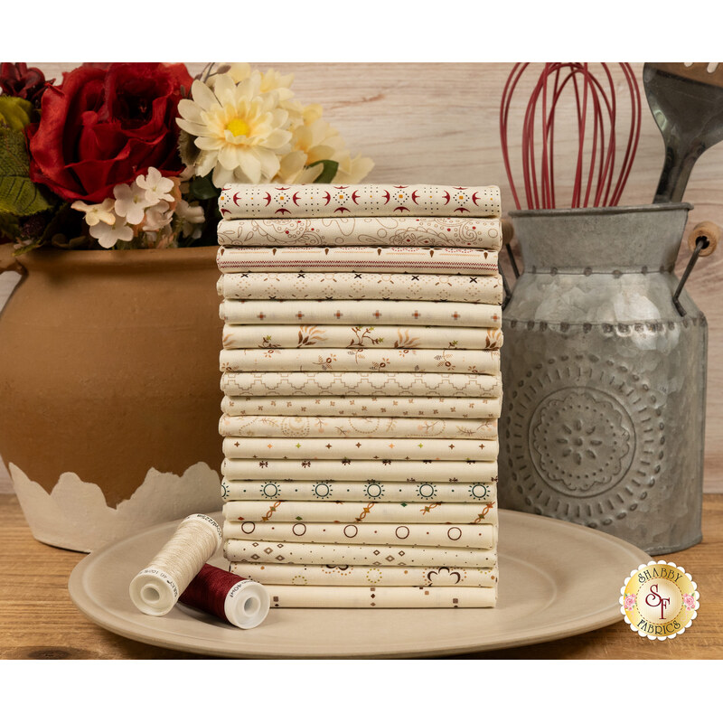 A stack of the fabrics included in the Buttercream & Brickle Bits FQ set, with a jug and a vase and a whisk and spatula and flowers and thread and a plate.
