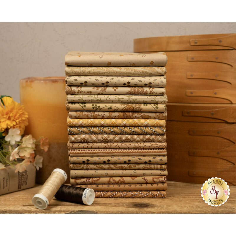 A stack of the nuetral fabrics included in the Toasted FQ Set, with boxes and a candle and flowers and a book and thread.