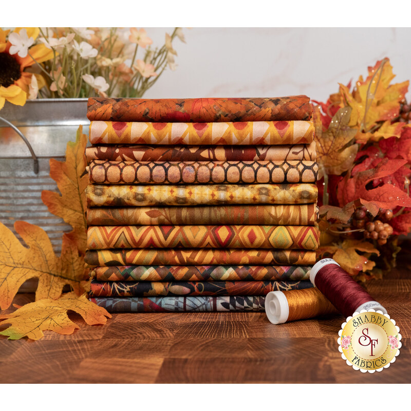 A stack of the fabrics included in the Falling Leaves collection, with leaves and a jug and flowers and thread.
