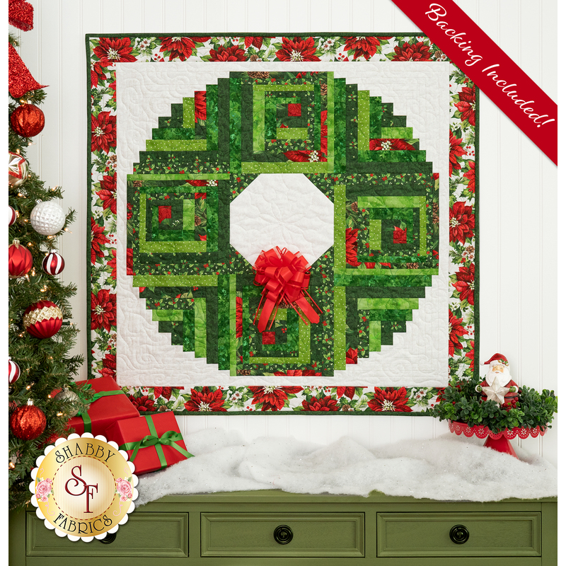 The completed Log Cabin Christmas Wreath in red, green, and white fabrics from the Yuletide Traditions collection, hung on a white paneled wall above a green dresser topped with a blanket of sparkly decorative snow, presents, and a Santa; a decorated Christmas tree peeks into frame from the left side.