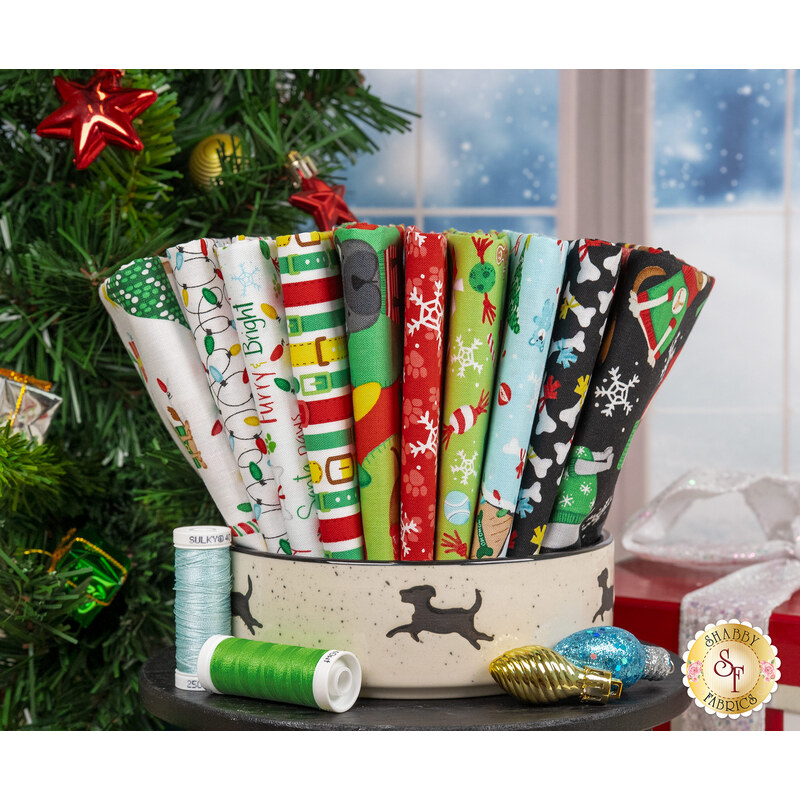 A stack of fabrics featured in the Merry Woof-Mas FQ set, with a dog bowl and a christmas tree and ornaments and presents and thread.