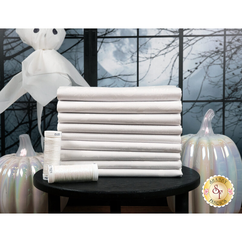 A stack of the white on white fabrics included in the Essentials Spooky Spectacle FQ set, with a moon and a window and a ghost and pumpkins and thread.