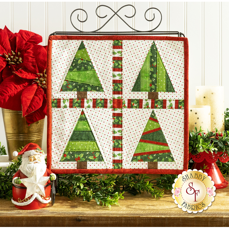 The completed Foundation Paper Piecing Series 2 project for December, an arrangement of four dynamic Christmas trees in red, green, and white fabrics, staged on a rustic wooden tabletop from a craft stand with coordinating decor.