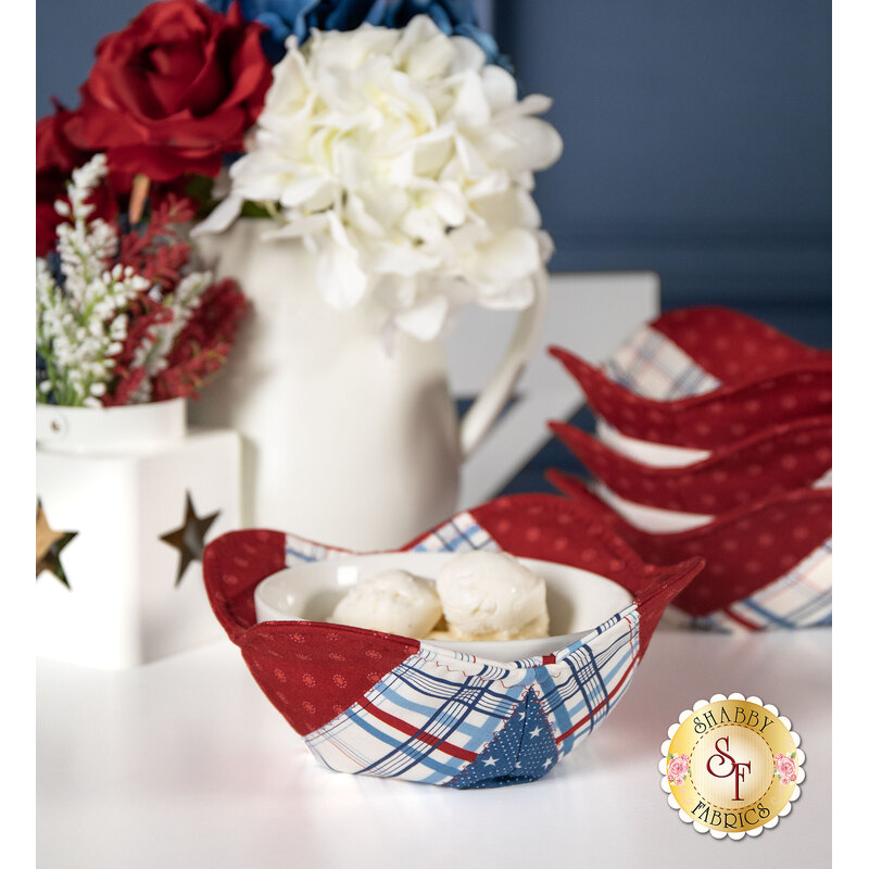 The completed bowl cozies, colored in patriotic fabrics from the Grand Haven collection, staged on a white table with coordinating flowers and decor. A blurred stack of bowl cozies sits in the background.