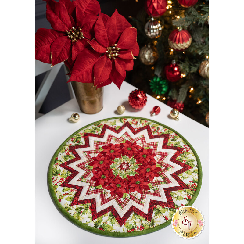 The completed Point of View Kaleidoscope Folded Star Table Topper colored in Holly Berry Park, displayed on a white table with coordinating flowers and Christmas decor.