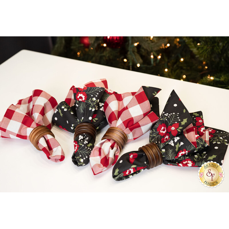 An angled shot of the four napkins slightly fanned out on a white table top. A decorated and lit Christmas tree can be seen over the edge of the table.