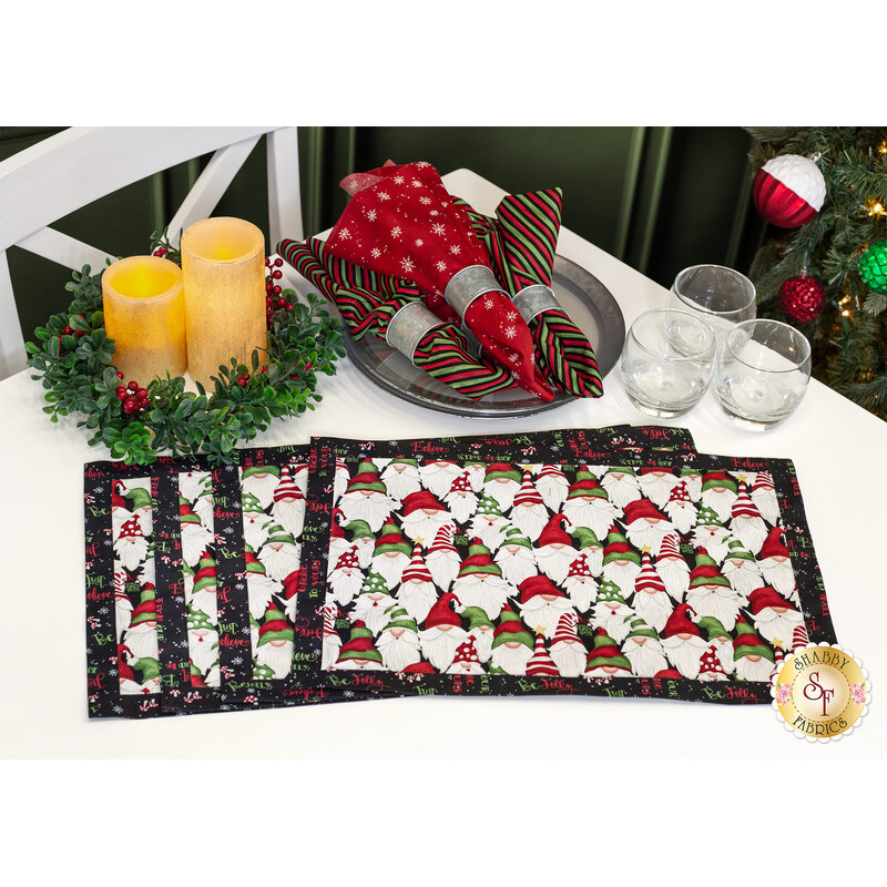 The four completed self binding placements, colored in the Our Gnome to Yours collection and staged on a white table with coordinating decor, glasses, and cloth napkins, with a decorated Christmas tree peeking into the frame.