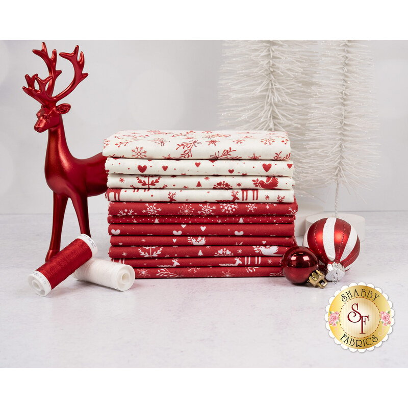 A stack of the fabrics featured in the Nordic Noel FQ set, with a Deer statue and trees and ornaments and thread.