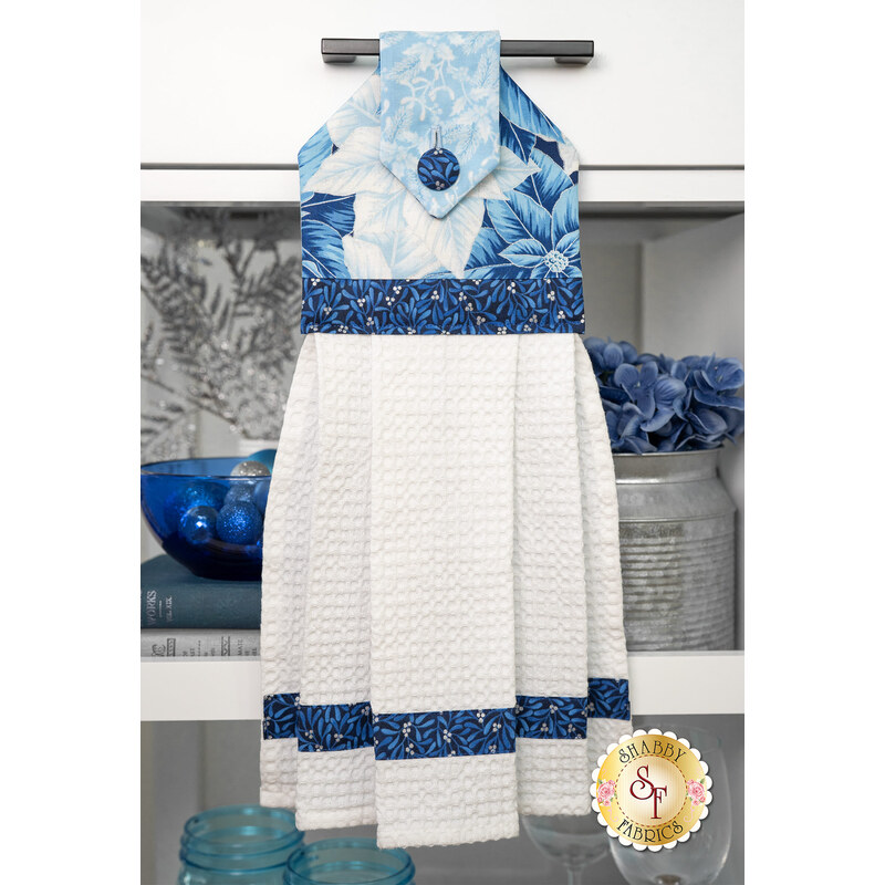 The completed poinsettia themed towel, colored in icy blue fabrics from the Holiday Flourish - Festive Finery collection, staged on a kitchen drawer with coordinating winter-themed decor in the background.