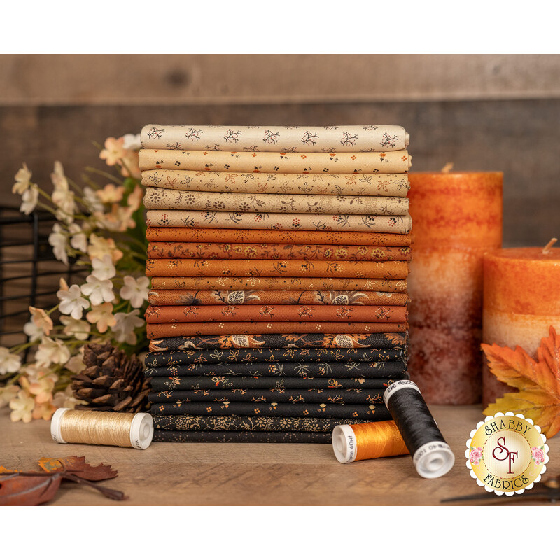 A stack of the fabrics inclided in the Cheddar & Coal III collection, with candles and flowers and leaves and pinecones and thread.