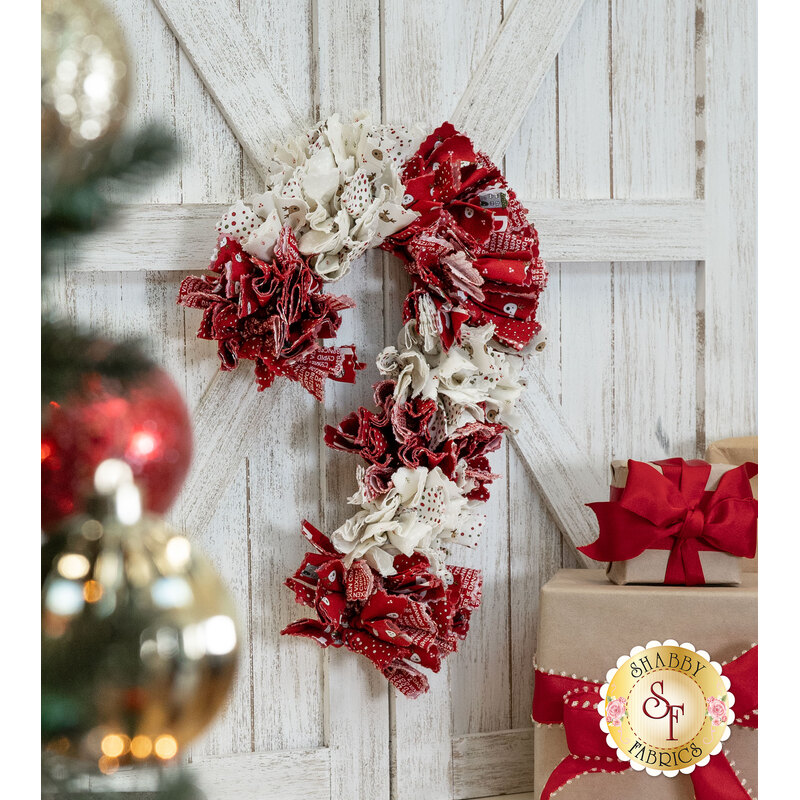 The completed Candy Cane Wreath, colored in red and cream fabrics from the On Dasher collection from Moda Fabrics, hung on a white wooden barn door beside craft wrapped presents. Red and golden baubles on a tree peek into frame from the left side.