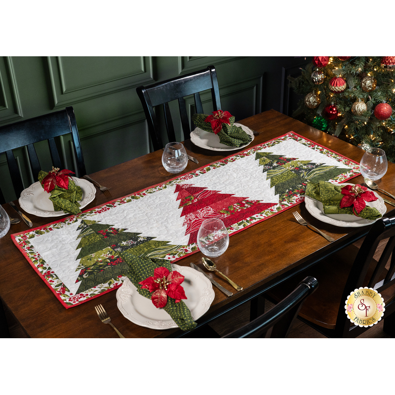 The completed Tree Farm Table Runner in Pine Valley fabrics, staged on a rustic wood table with coordinating tableware, glasses, napkins, and a decorated Christmas tree.