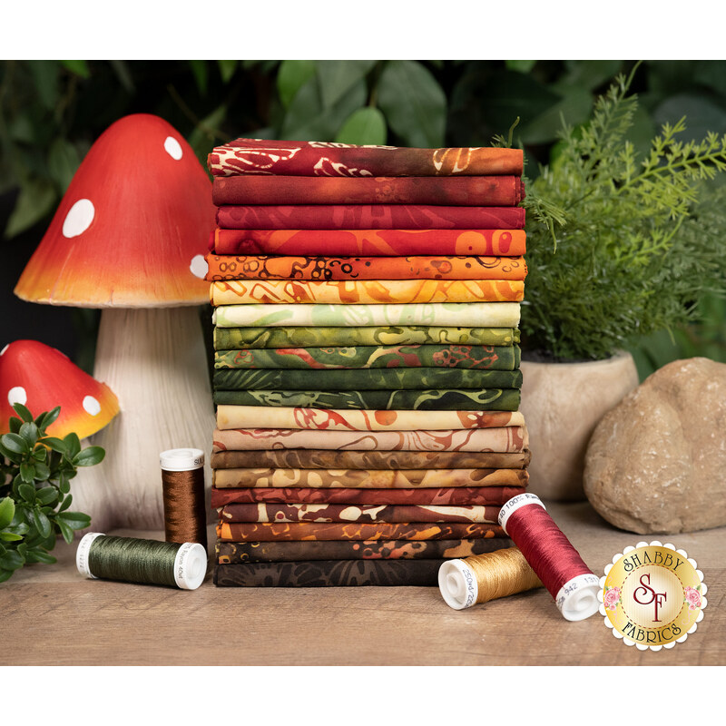 A stack of the fabrics from the Forest Flora collection, with a mushroom and rocks and plants and thread.