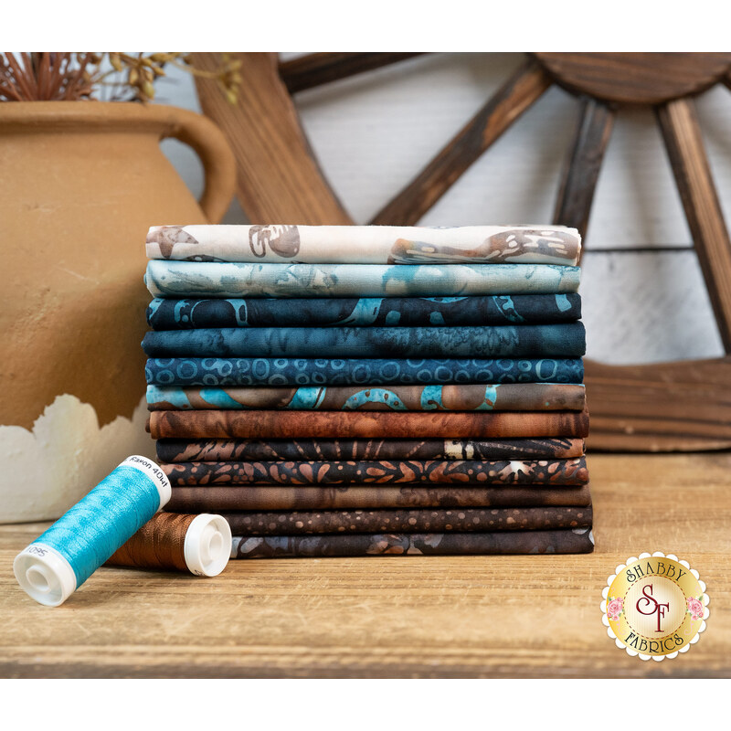 A stack of fabrics included in the On the Range collection, with a wagon wheel and a vase and plants and thread.