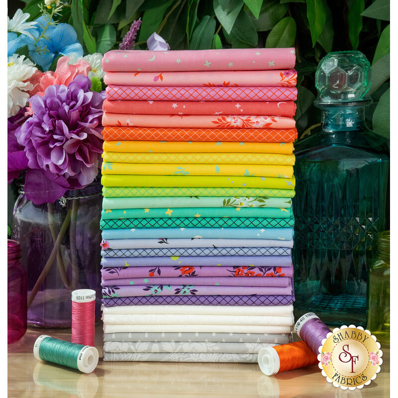 A stack of the fabrics from Tula's True Colors collection for 2025, with a decanter and jars and flowers and thread.