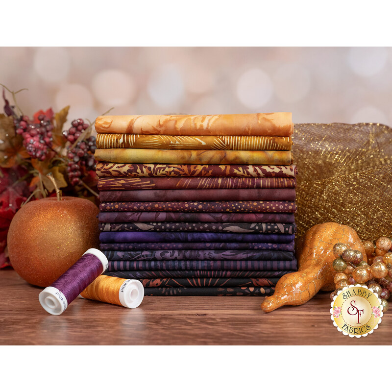 A stack of the fabrics included in the Harvest Glow collection, with leaves and fruit and a squash and thread