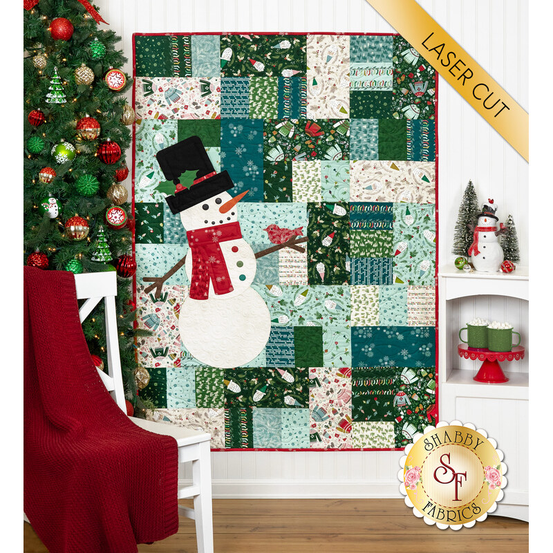 A festive quilt featuring a snowman with a patchwork design, hanging beside a decorated Christmas tree and a chair.