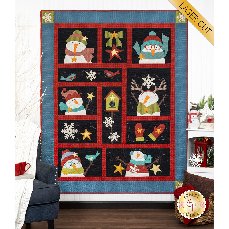 The completed Chilling With My Snowmies quilt colored in red, black, and blue Stitcher's Flannel fabrics, hung on a white paneled wall and bordered by coordinating winter decor.