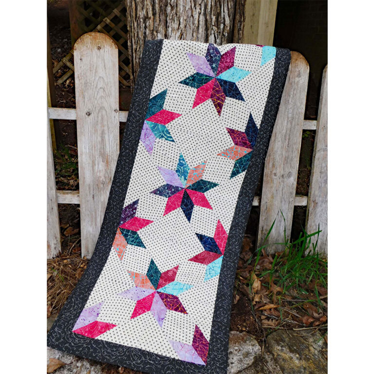 A colorful quilt with star patterns in shades of blue, pink, and purple, laid against a wooden fence.