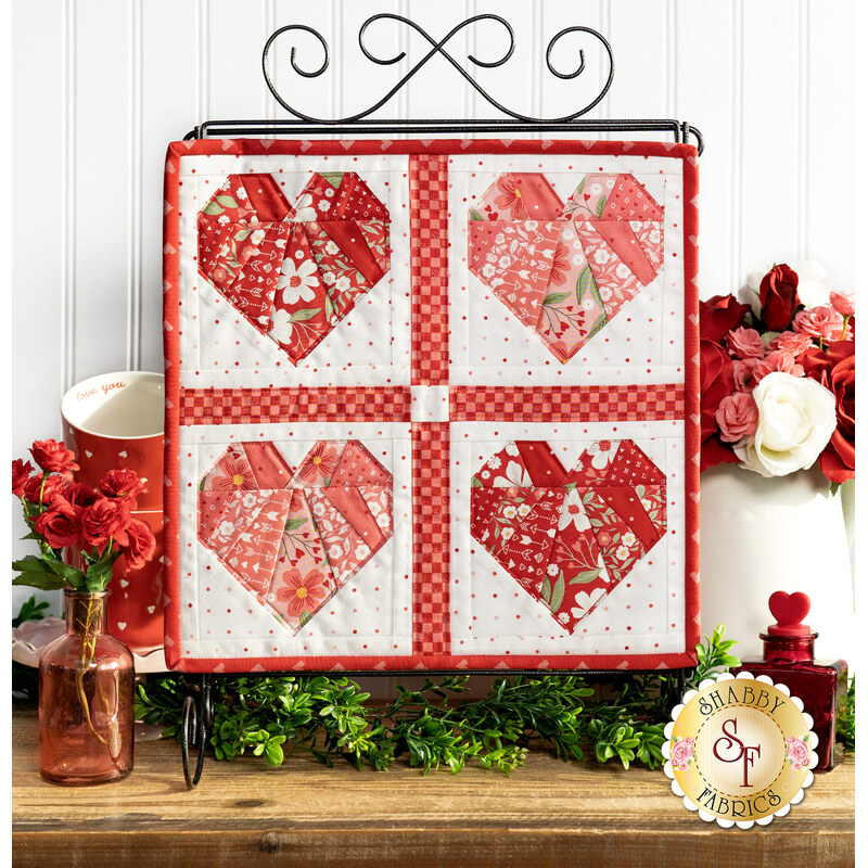 The completed Foundation Paper Piecing Series 2 project for February, an arrangement of four hearts in red, pink, and white fabrics, staged on a rustic wooden tabletop from a craft stand with coordinating decor.