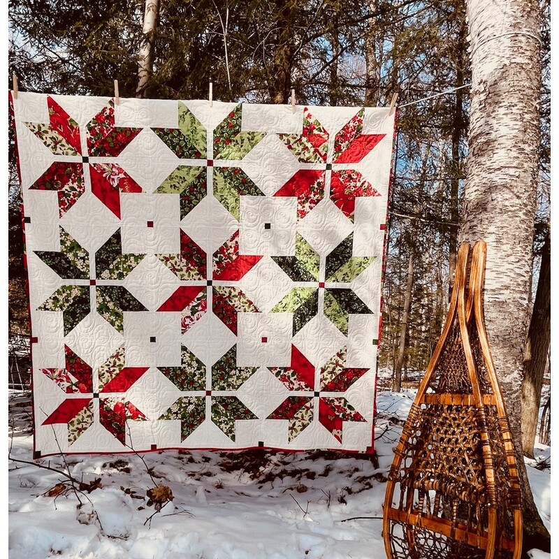 A head on shot of the completed Vintage Stars quilt, colored in cream, red, and green fabrics, hung on a clothesline and staged outdoors in a snowy wood with snowshoes.