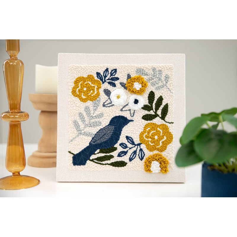 A decorative punch needle piece featuring a blue bird among yellow and white yarn flowers on a cream background.