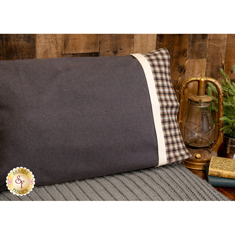 An angled shot of the Magic Pillowcase in gray, staged on a bed with a matching blanket in a cabin setting. A gold lantern and a stack of books sit on a table next to the bed.