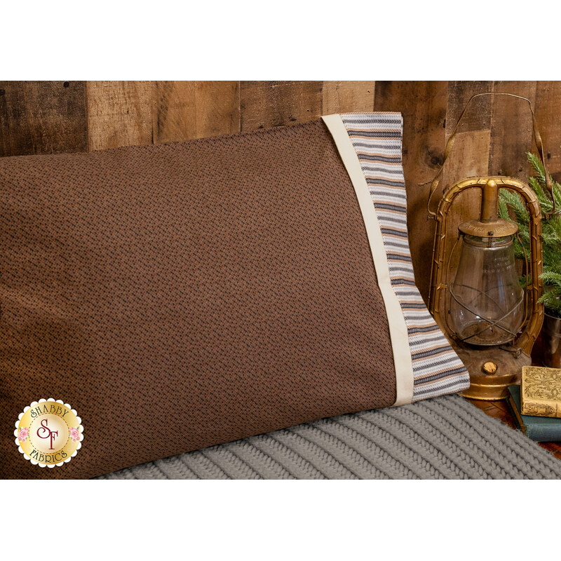 An angled shot of the Magic Pillowcase in brown, staged on a bed with a matching blanket in a cabin setting. A gold lantern and a stack of books sit on a table next to the bed.