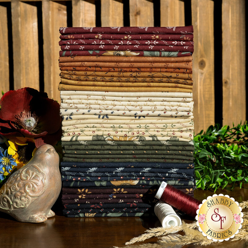 A stack of the 40 fabric swatches in earthy tones with various patterns included in Wheatland by Moda Fabrics, with flowers and thread and wheat stalks and a bird statue.