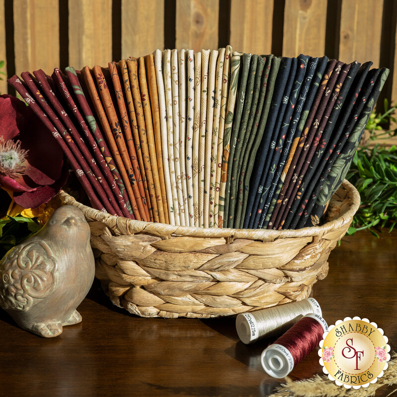 A stack of the 40 fabric swatches in earthy tones with various patterns included in the Wheatland F8 set by Moda Fabrics, with flowers and thread and wheat stalks and a bird statue.