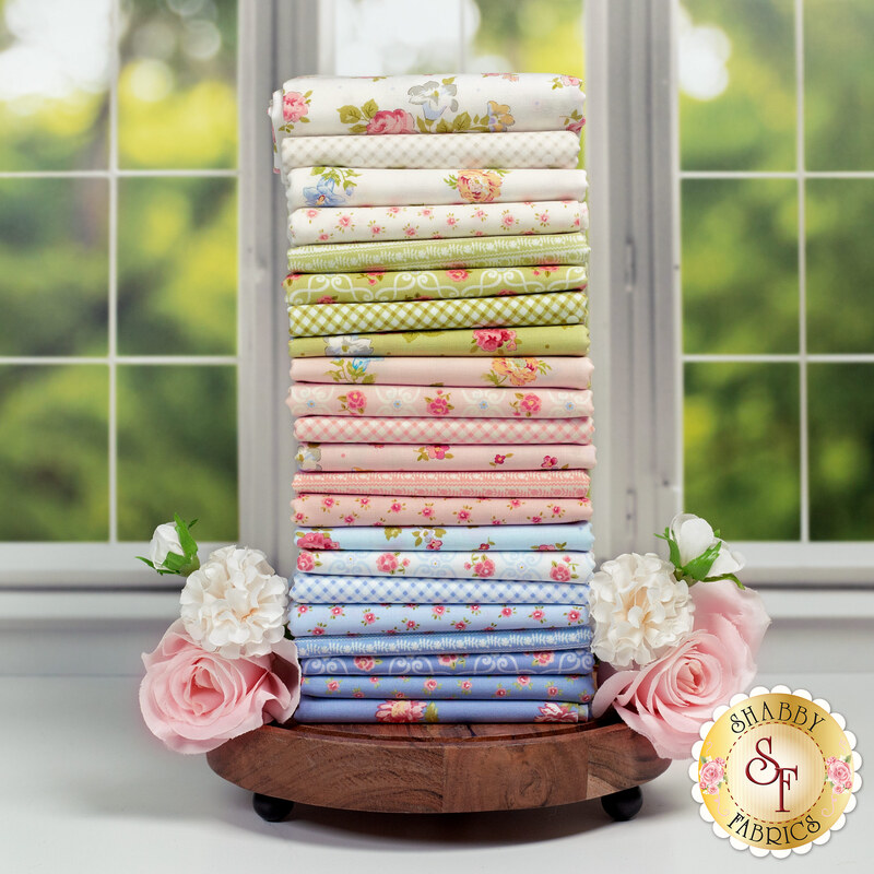 A stack of the fabric set Memory Lane by Moda Fabrics, featuring floral designs and pastel colors, with flowers and a wooden stand and a window.