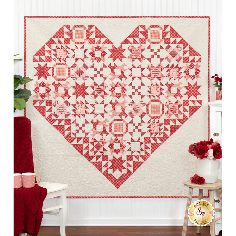 A large heart-shaped quilt in red and cream patterns hangs on a white wall, surrounded by flowers.