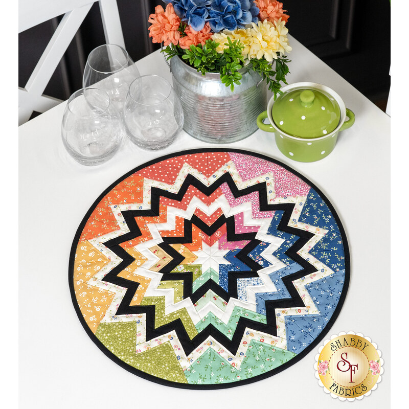 A top down shot of the finished Folded Star Table Topper colored in rainbow fabrics from the Cali & Co collection by Moda Fabrics. It sits on a white table, staged with glassware and coordinating flowers.