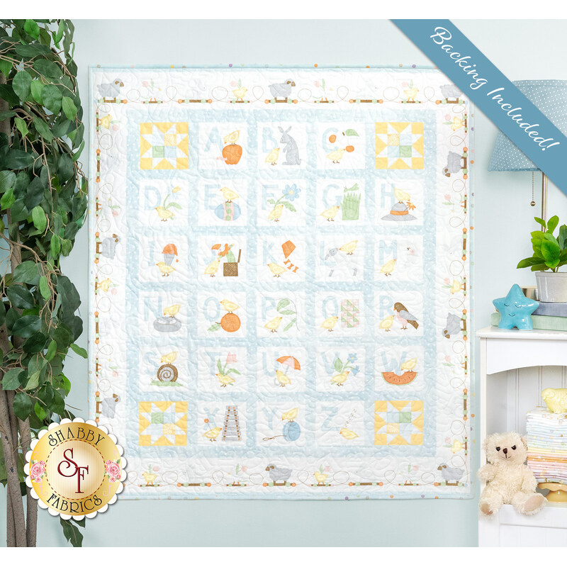 A childrens quilt featuring alphabet blocks with various whimsical patterns hangs on a wall, surrounded by plants and decor.