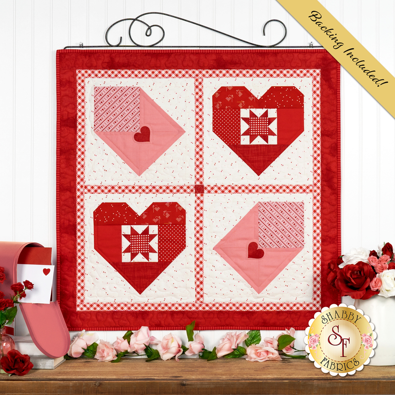 The completed Pieceful Patchwork February Wall Hanging, a Valentine's Day themed project with pieced hearts and love letters, colored in red, white, and pink fabrics, hung from a craft hanger on a white paneled wall, and staged with coordinating decor.