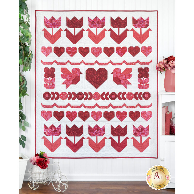 A patchwork quilt featuring red and pink hearts, flowers, and birds arranged in a pattern on a white background surrounded by coordinating decor.