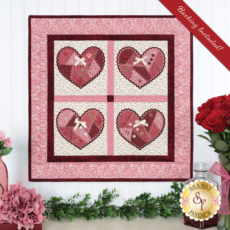 A quilt depicting four heart patterns in pink and red, framed by a floral design, with embellishments.