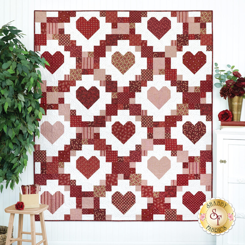 A quilt featuring a repeating pattern of hearts in shades of red and pink, displayed on a white wall next to coordinating decor. 