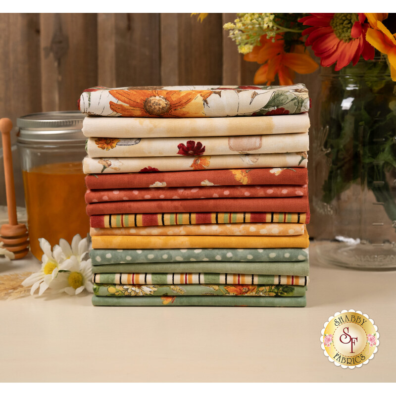 A stack of the fabrics featured in the Harvest Blooms FQ set, with flowers and jars and honey and a honey wand and wheat stalks and thread.