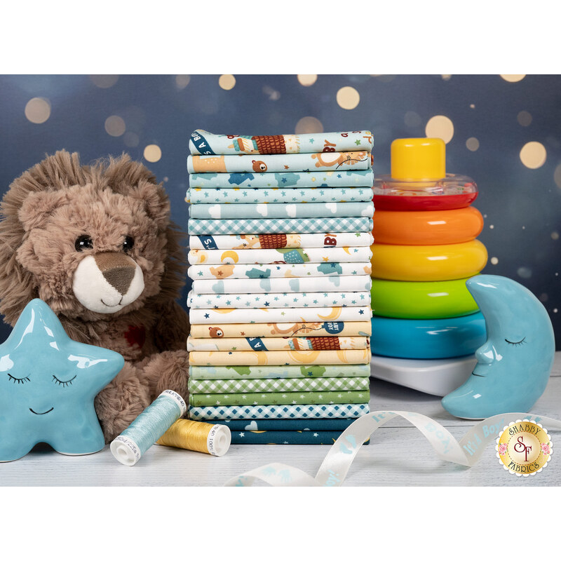 A stack of the fabrics in the Baby Boy collection, with ribbon and a star and a moon and a lion plush and a stacking ring toy and thread. 