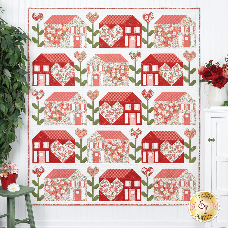 The completed Love Blooms at Home Quilt, colored in white, pink, and red fabrics from Moda's Love Blooms collection, hung on a white paneled wall and staged with coordinating decor and flowers.