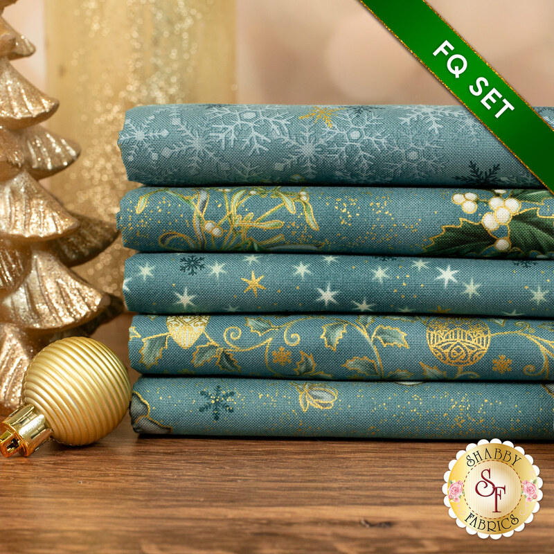 A stack of the five fabrics included in the 5 Fat Quarter Set Teal/Gold by Stof Fabrics, with candles and an ornament.