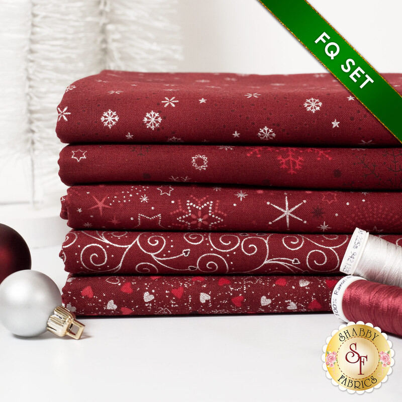 A stack of the five fabrics included in the 5 Fat Quarter Set Red/Silver by Stof Fabrics, with ornaments and thread.