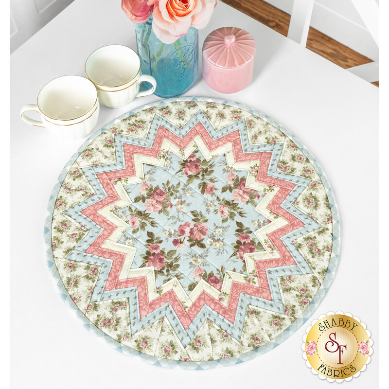 The completed Point of View Folded Star Table Topper, colored in soft pink, blue, and cream fabrics. The topper is staged on a white table with coordinating florals and cups.