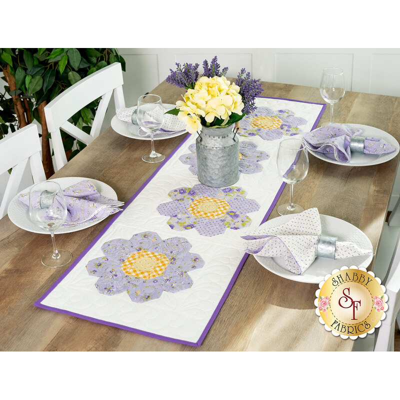 The table runner displayed on a wooden table that's set with a small milk can holding flowers and white porcelain plates set with coordinating cloth napkins, tableware, and wine glasses.