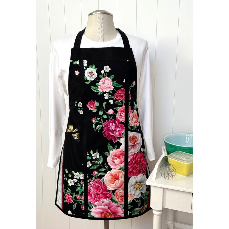 A floral-patterned black apron displayed on a mannequin, paired with kitchen utensils and bowls.