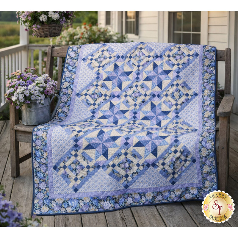 The finished Morning Blooms Patchwork Quilt in warm blues, purples, and creams, draped over a wooden bench on a porch surrounded by coordinating décor.  