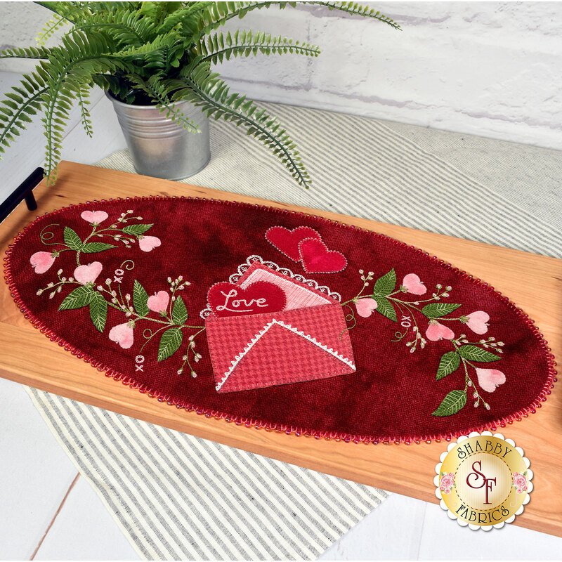 The completed PS I Love You wool mat, colored in vibrant red, pink, and green wool and embroidery thread, staged on a wooden tray with black handles and a green fern in a silver planter.