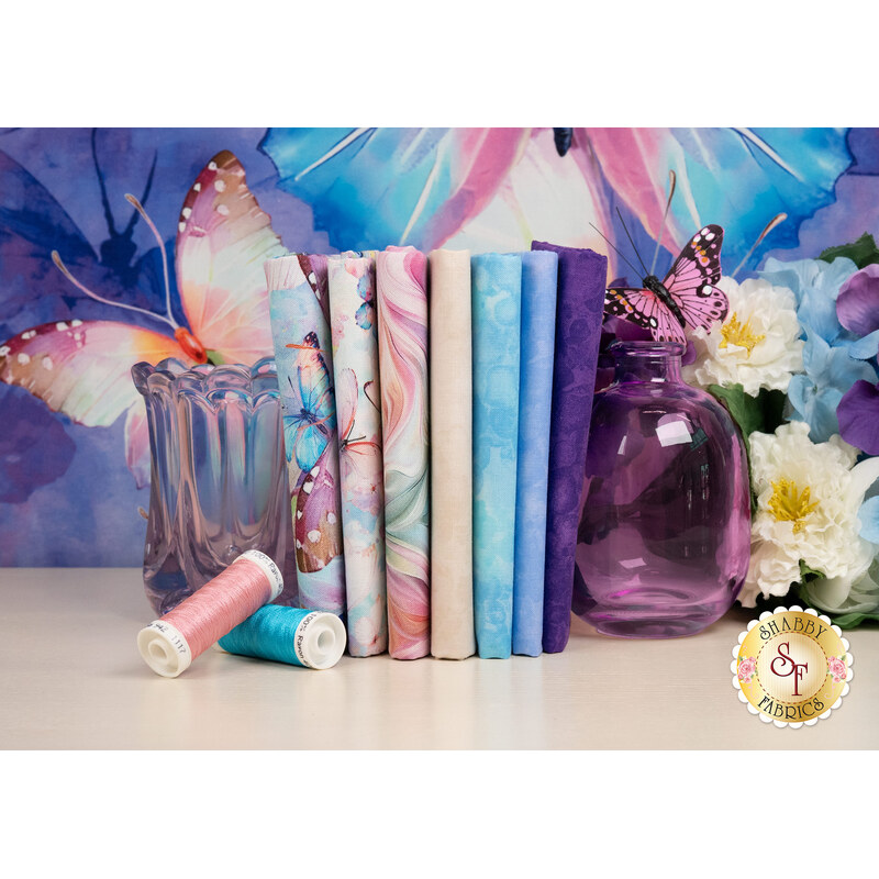 A stack of the fabrics included in the Flutter FQ set, with the panel in the background and vases and flowers and a butterfly and thread.